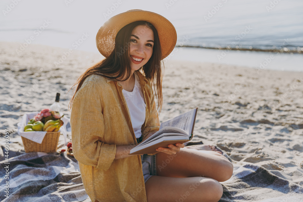 Full length young happy traveler tourist woman in straw hat shirt summer clothes reading book sit on plaid have picnic outdoors on sea sand beach background People vacation lifestyle journey concept