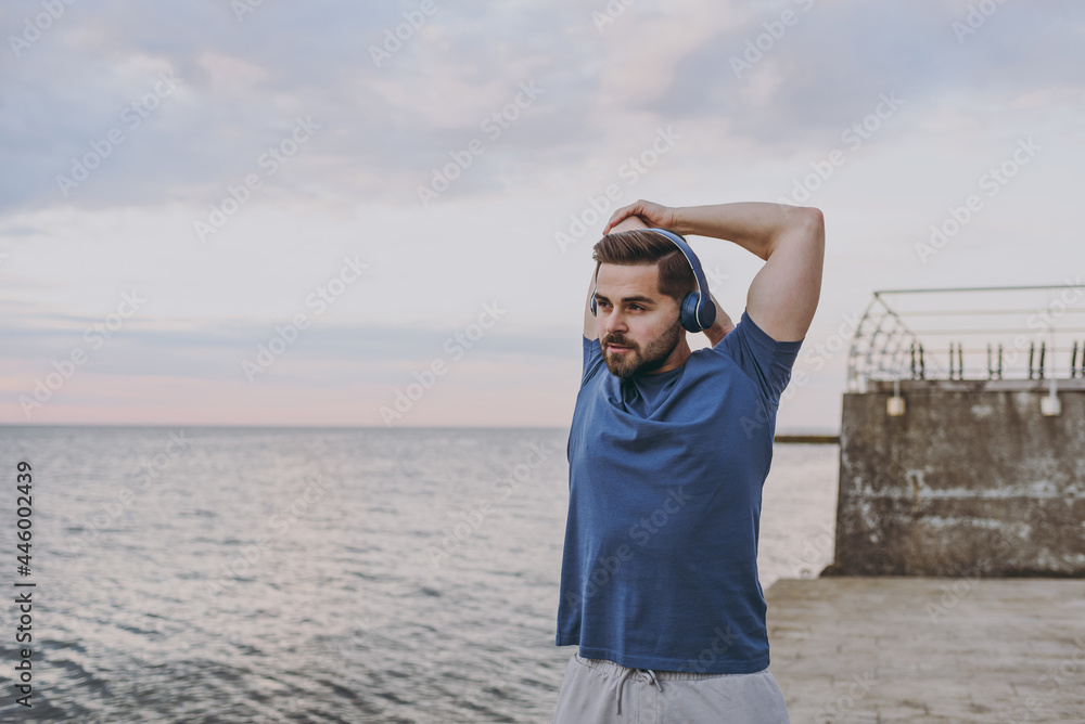 Young strong sporty athletic toned fit sportsman man in sports clothes headphones warm up training do hand exercises at sunrise sun dawn over sea beach outdoor on pier seaside in summer day morning Young strong sporty athletic toned fit sportsman man in sports clothes headphones warm up training do hand exercises at sunrise sun dawn over sea beach outdoor on pier seaside in summer day morning