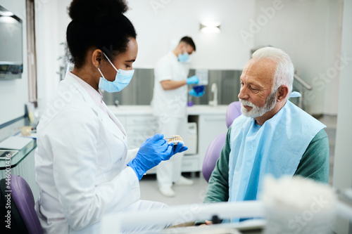 Photos African American dentist using artificial dentures while communicating with senior patient at dentist's office