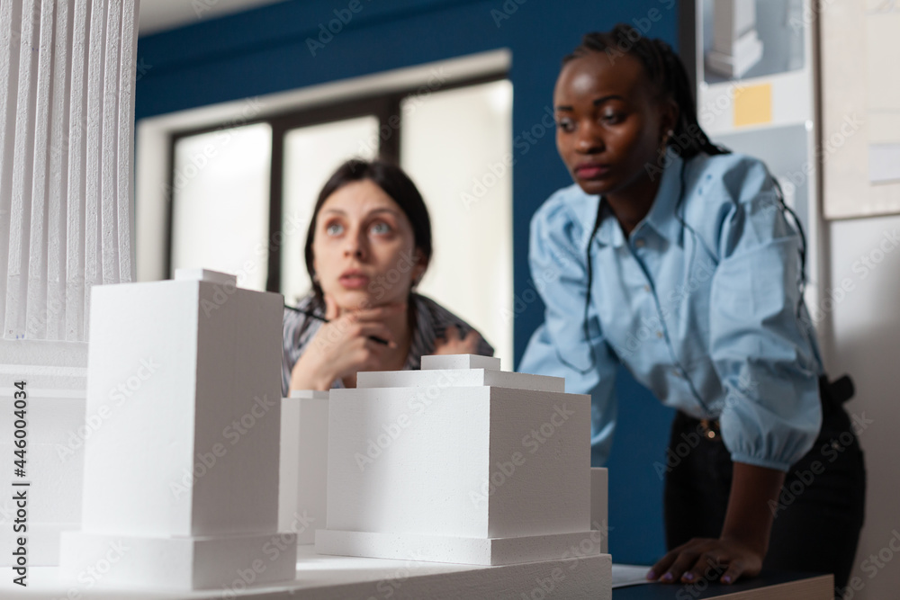 Multi ethnic diverse team of architects at office working on building model maquette blueprint plans for occupation. Professional women looking at modern layout construction design Multi ethnic diverse team of architects at office working on building model maquette blueprint plans for occupation. Professional women looking at modern layout construction design