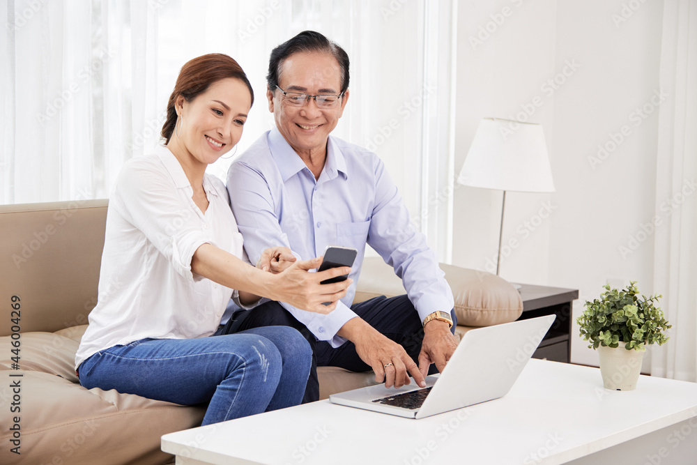 Smiling senior couple sitting on sofa in living room and taking selfie on smartphone or using mobile application for video calling family and friends Smiling senior couple sitting on sofa in living room and taking selfie on smartphone or using mobile application for video calling family and friends