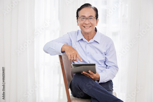 Positive elderly man sitting on chair and reading news or checking social media on tablet computer