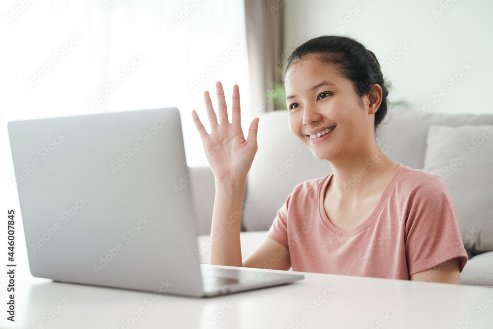 Young Asian woman using laptop computer for online video conference call waving hand making hello gesture in the living room. Young Asian woman using laptop computer for online video conference call waving hand making hello gesture in the living room.