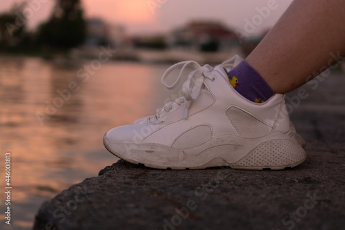 Legs in white sneakers on the bank of the river at sunset