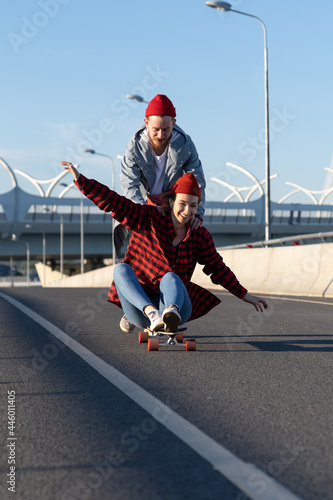 Happy couple of lovers chilling outdoors relaxing ride longboard, laugh and have fun together. Carefree young trendy woman sit on long board skate with hipster man pushing her back to slide city road
