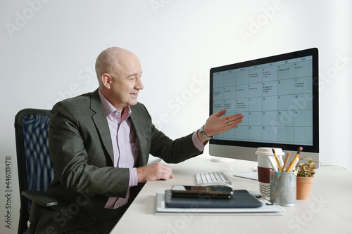 Mature businessman in suit sitting at the table and pointing at computer monitor, he working with graphics