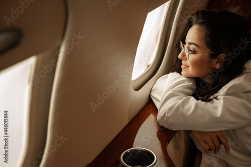 Young beautiful brunette girl in a white suit flies in a charter plane on business admires the scenery from the porthole