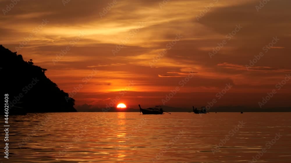 Classical view of tropical sunset, half of sun seen at horizon, dark and colorful scenery. Calm sea water at shallow Lohdalum Bay, long tail boats stay at distance, mountain silhouette at left Classical view of tropical sunset, half of sun seen at horizon, dark and colorful scenery. Calm sea water at shallow Lohdalum Bay, long tail boats stay at distance, mountain silhouette at left
