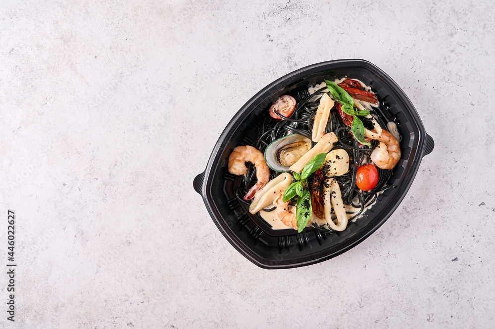 Black spaghetti Nero with seafood, tomatoes and basil in black plastic container on light background. Designer food concept for take away or delivery