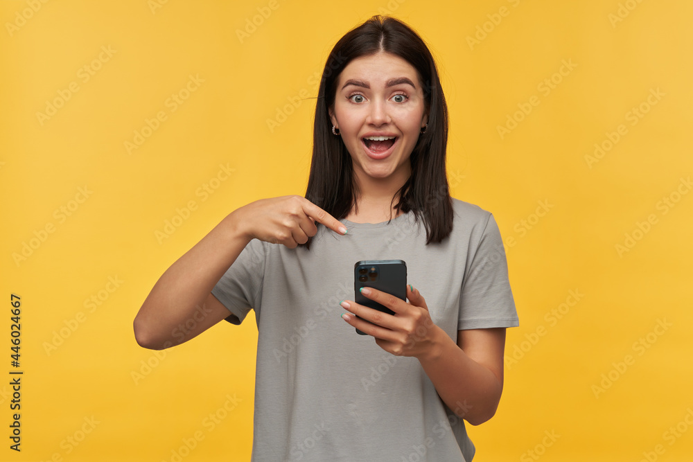 Happy excited young woman with dark hair and opened mouth in gray tshirt looks amazed using and pointing at mobile phone over yellow background Happy excited young woman with dark hair and opened mouth in gray tshirt looks amazed using and pointing at mobile phone over yellow background