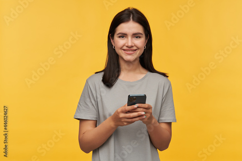 Cheerful attractive young woman with dark hair in gray tshirt using smartphone and looking at camera over yellow background