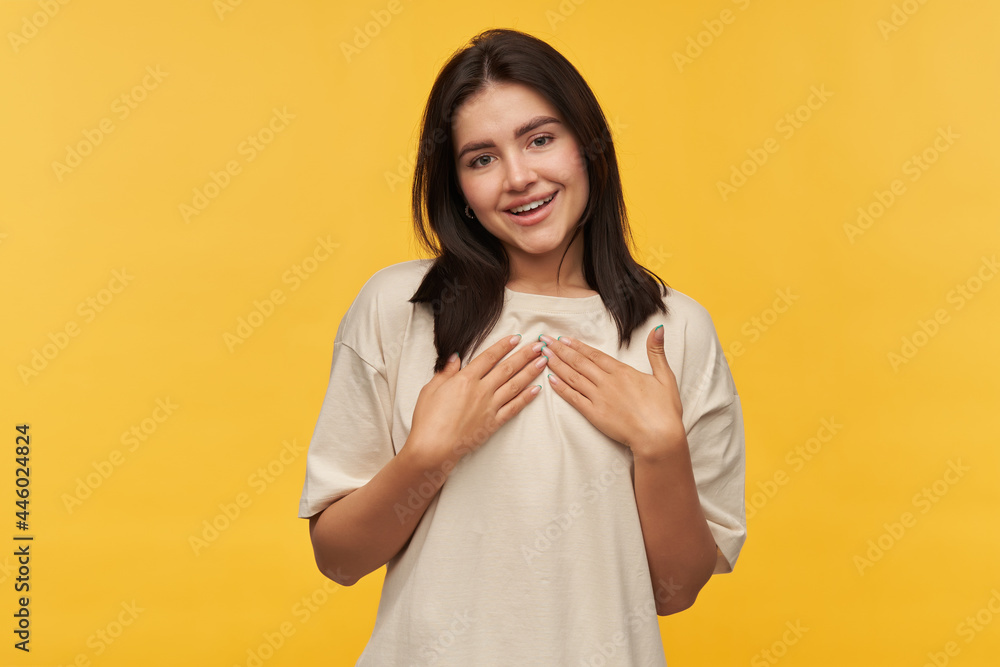Smiling beautiful young woman with dark hair in white tshirt keeps hands on chest and showing grateful gesture over yellow background Smiling beautiful young woman with dark hair in white tshirt keeps hands on chest and showing grateful gesture over yellow background
