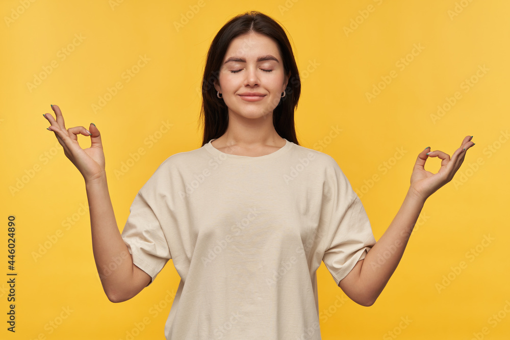Calm relaxed young woman with dark hair in white tshirt keeps hands in mudra zen gesture and meditating over yellow background