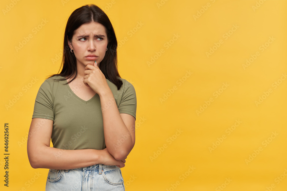 Sad unhappy brunette young woman in casual clothes keeps hands folded and looking away to the side at copyspace over yellow background Sad unhappy brunette young woman in casual clothes keeps hands folded and looking away to the side at copyspace over yellow background