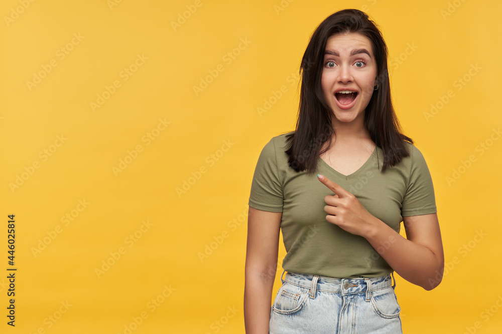 Surprised beautiful young woman with dark hair and opened mouth in casual clothes pointing away to the side at empty space over yellow background Surprised beautiful young woman with dark hair and opened mouth in casual clothes pointing away to the side at empty space over yellow background