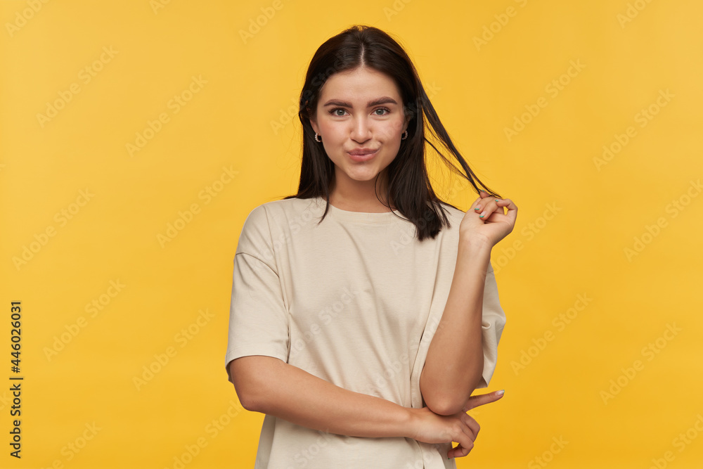 Obraz premium Studio portrait of smiling attractive young woman in white tshirt standing and touching her dark hair over yellow background Looking at camera