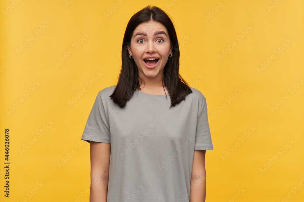 Happy amazed young woman with dark hair and opened mouth in gray tshirt looks excited and looking at camera over yellow background Happy amazed young woman with dark hair and opened mouth in gray tshirt looks excited and looking at camera over yellow background
