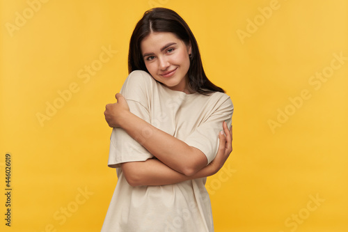 Portrait of cute charming brunette young woman in white tshirt standing and hugging herslf by hands over yellow background Looking at camera © timtimphoto