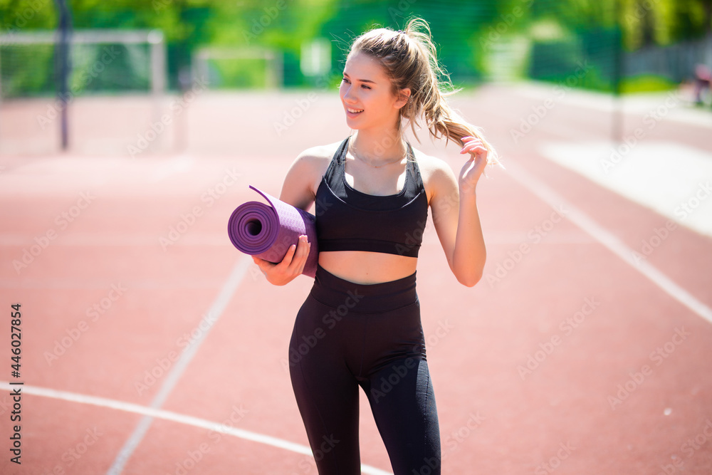 Fitness girl with yoga mat standing outdoor in nature. Fit woman with exercise accessory at stadium. Fitness girl with yoga mat standing outdoor in nature. Fit woman with exercise accessory at stadium.