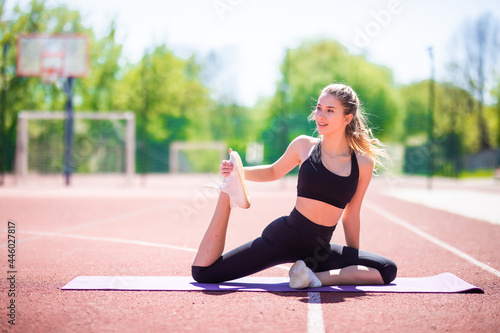 Sport woman in sportswear doing stretching one leg while sitting on mat on stadium track with red coating at sunny day © F8  \ Suport Ukraine