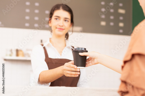 Takeaway coffee cup in two hands of customer and beautiful caucasian barista woman who looks at other customers, focusing at coffee cup