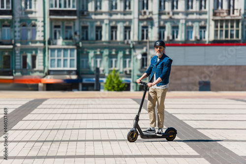 full length of middle aged man in sunglasses and helmet riding e-scooter in urban city