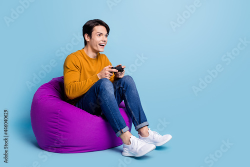 Full length photo of impressed brunet young guy sit on bag playstation wear sweater jeans isolated on blue color background