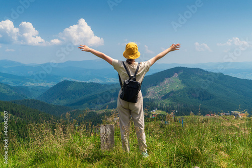 A girl traveler with a backpack stands with her arms outstretched in the mountains in summer. The top of the mountain evokes good or uplifting mood