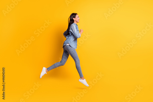 Full size profile photo of cool optimistic brunette lady go run wear blue shirt jeans sneakers isolated on yellow color background