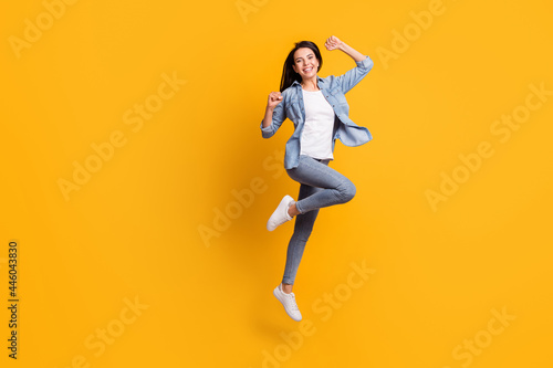 Full size profile photo of cool hooray brunette lady run jump wear blue shirt jeans sneakers isolated on yellow background