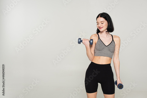 Cheerful sportswoman with vitiligo working out with dumbbells isolated on grey