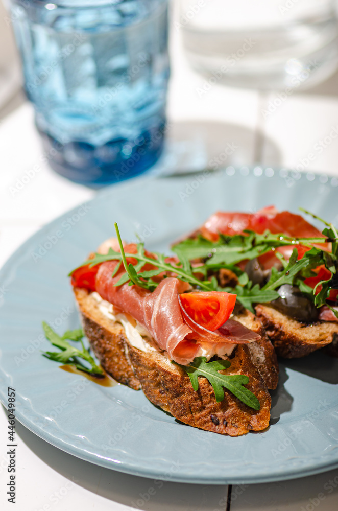 Bruschetta with dark baguette, jamon, arugula and tomato on a white tile table, bright hard sunlight.