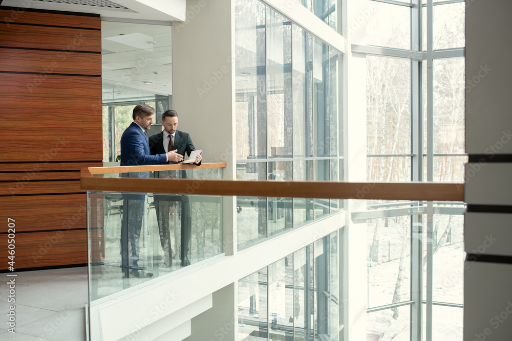Two businessmen discussing document together while standing in the corridor of the modern office building Two businessmen discussing document together while standing in the corridor of the modern office building