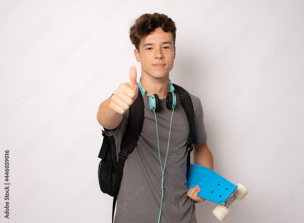 Schoolboy in headphones holding skateboard with thumb up isolated over white background. Schoolboy in headphones holding skateboard with thumb up isolated over white background.