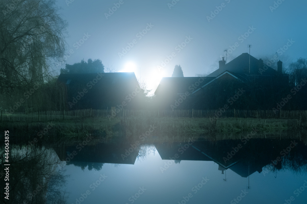 Houses silhouetted against bright street lights. reflected in water. On a moody, misty, winters night. UK