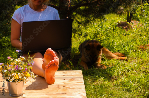 Remote work. Woman works with laptop in a green garden. Pine and dog on the background.