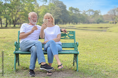 senior caucasian grandfather and grandmother sitting in park while using video call from smart phone to grandchildren with happy face and laughing