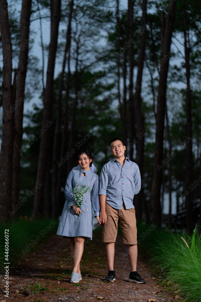 Portrait Pre Wedding of an Asian couple dressed in the same blue tones posing in a pine garden expressing their love in happiness behind an out-focused pine tree.