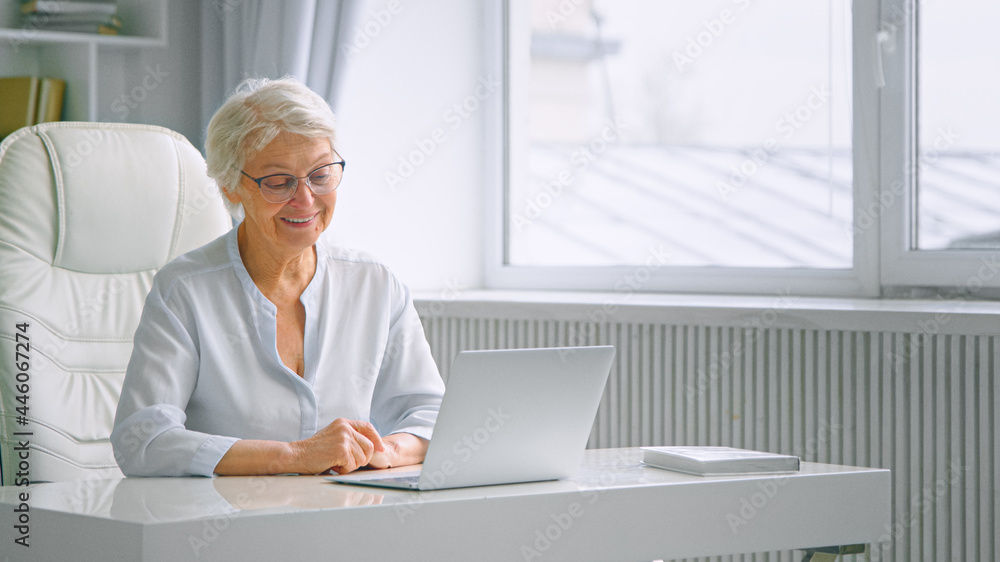 Old company woman worker waves hand greeting colleagues at online meeting and sitting at white table Old company woman worker waves hand greeting colleagues at online meeting and sitting at white table