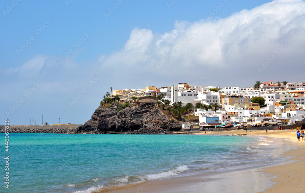 View of part of the town of Morro Jable with white buildings on a rocky shore and the beach with turquoise blue ocean water. Fuerteventra, Canary Islands, Spain