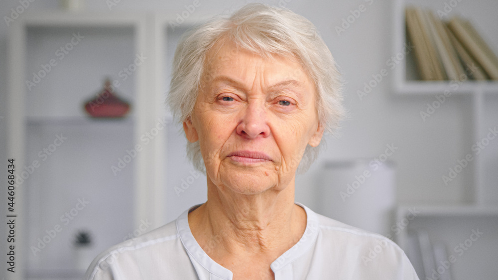 Old company lady manager with short hair looks straight standing against blurred white bookshelves Old company lady manager with short hair looks straight standing against blurred white bookshelves