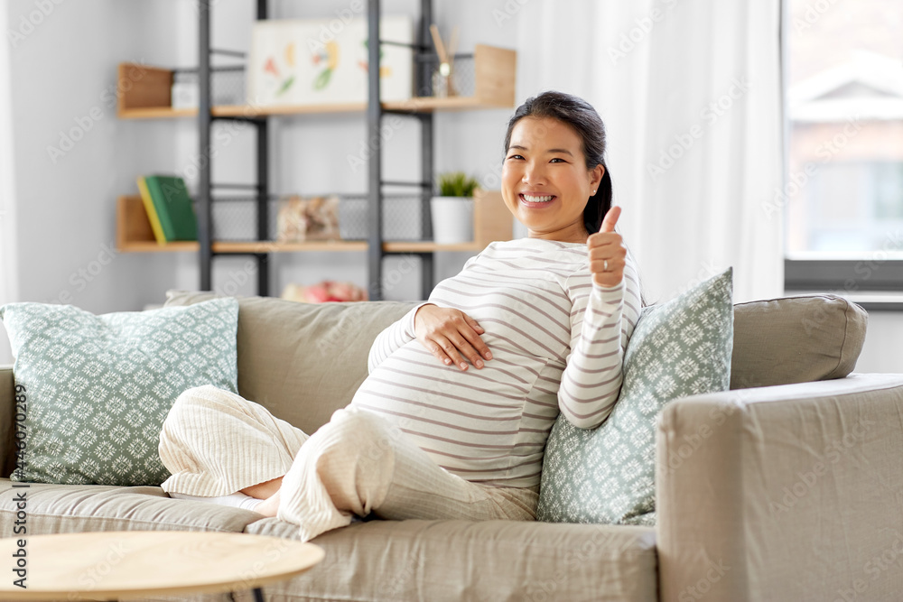 pregnancy, rest, people and expectation concept - happy smiling pregnant asian woman sitting on sofa at home and showing thumbs up pregnancy, rest, people and expectation concept - happy smiling pregnant asian woman sitting on sofa at home and showing thumbs up