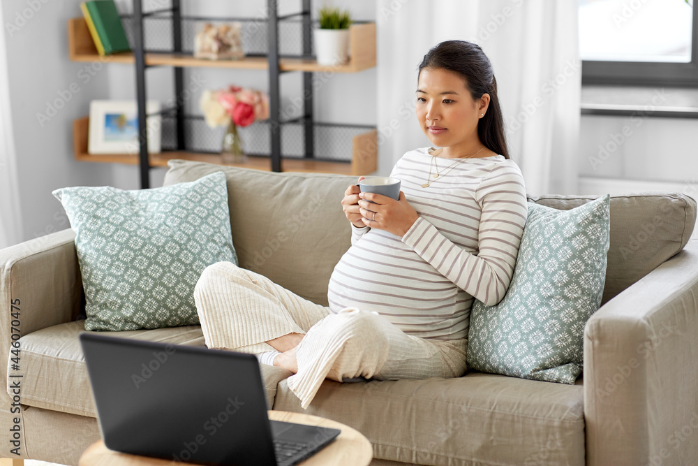 pregnancy, rest, people and expectation concept - pregnant asian woman with laptop computer sitting on sofa at home and drinking tea