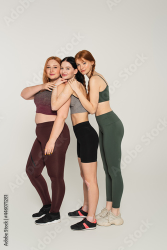 Full length of smiling body positive women in sportswear embracing each other on grey background