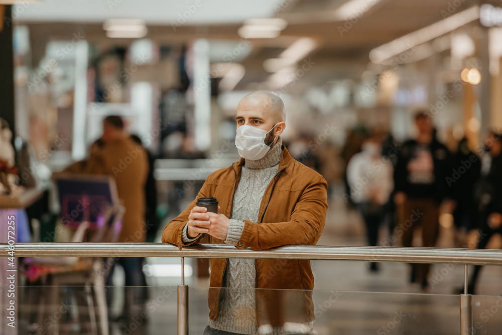 A man in a face mask to avoid the spread of coronavirus is holding a cup of coffee while waiting in the shopping center. A bald guy in a surgical mask is keeping social distance.