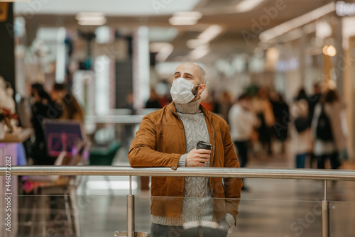 A man in a face mask to avoid the spread of coronavirus is holding a cup of coffee while waiting in the shopping center. A bald guy in a surgical mask is keeping social distance.