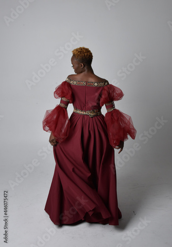 Full length portrait of pretty African woman wearing long red renaissance medieval fantasy gown  standing pose on a light grey studio background.