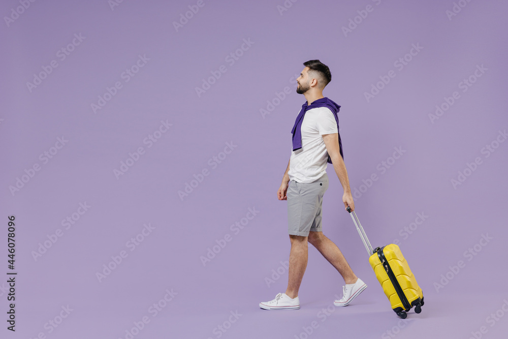 Full size body length side view fun traveler tourist young brunet man 20s wear white t-shirt purple shirt go move stroll hold yellow suitcase bag isolated on pastel violet background studio portrait. Full size body length side view fun traveler tourist young brunet man 20s wear white t-shirt purple shirt go move stroll hold yellow suitcase bag isolated on pastel violet background studio portrait.