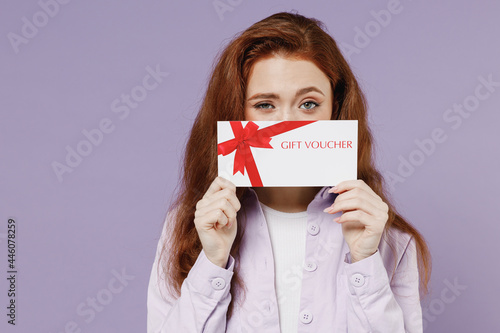 Happy young redhead curly woman 20s wears white T-shirt violet jacket hold hide behind gift certificate coupon voucher card for store isolated on pastel purple color wall background studio portrait