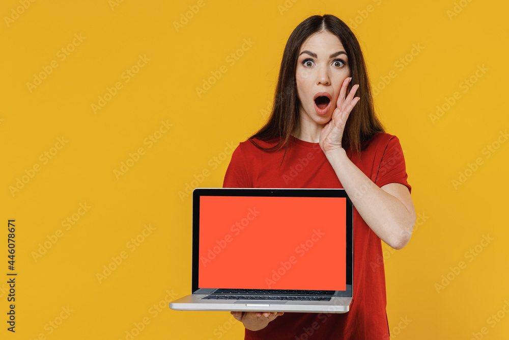 Naklejka premium Cute young brunette woman 20s wears basic red t-shirt hold use work on laptop pc computer with blank screen workspace area scream with hands near mouth isolated on yellow background studio portrait.